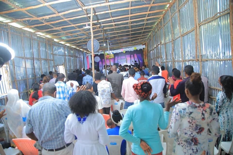 About 200 hundred people inside the church at Awasa at a worship service. The walls and roof are made of sticks and corrugated meta. The stage has a purple curtain behind it.
