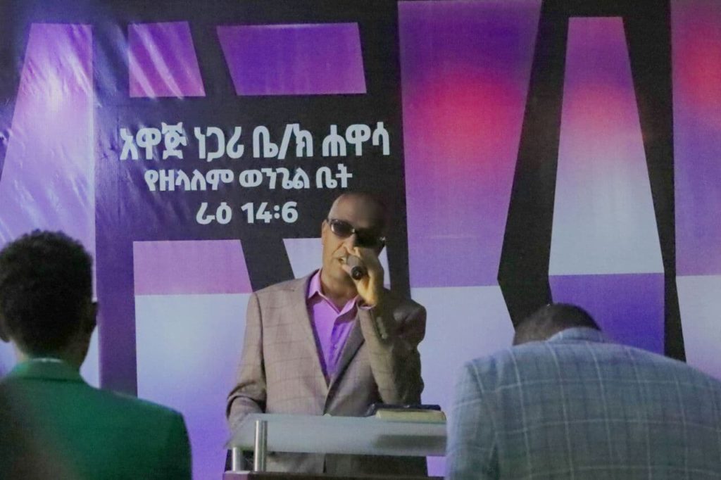 Berhanu speaking at the church in Awassa. He wears a gray suit and dark sunglasses.